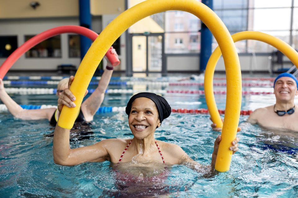 Several smiling people in a pool use pool noodles for exercise. The focus is on a woman in a swimsuit and swim cap, holding a yellow noodle overhead