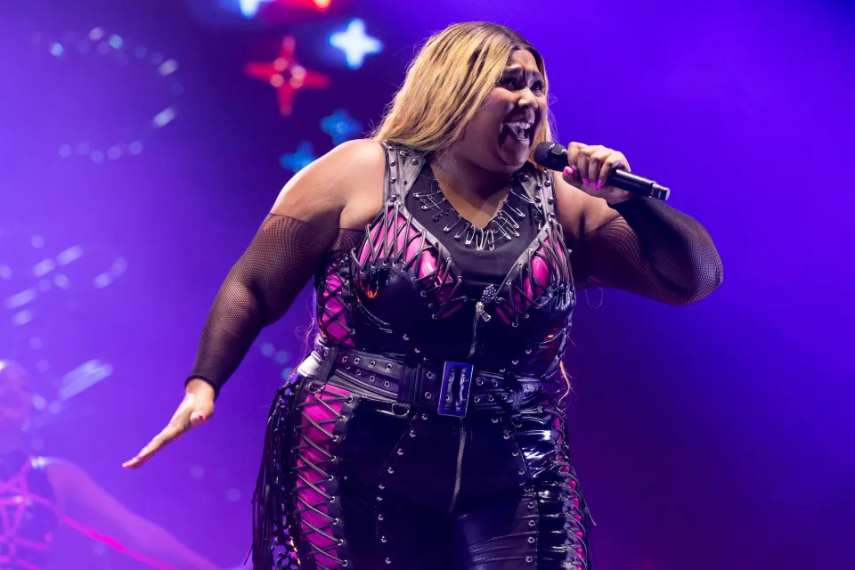 Lizzo singing into a microphone while performing on stage and wearing a black and pink leather suit.