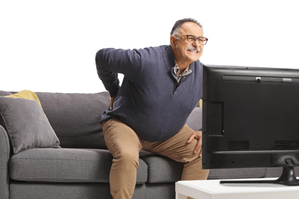 A man is sitting on a couch, holding his lower back in pain, while watching television. The photo implies he may have back discomfort or strain