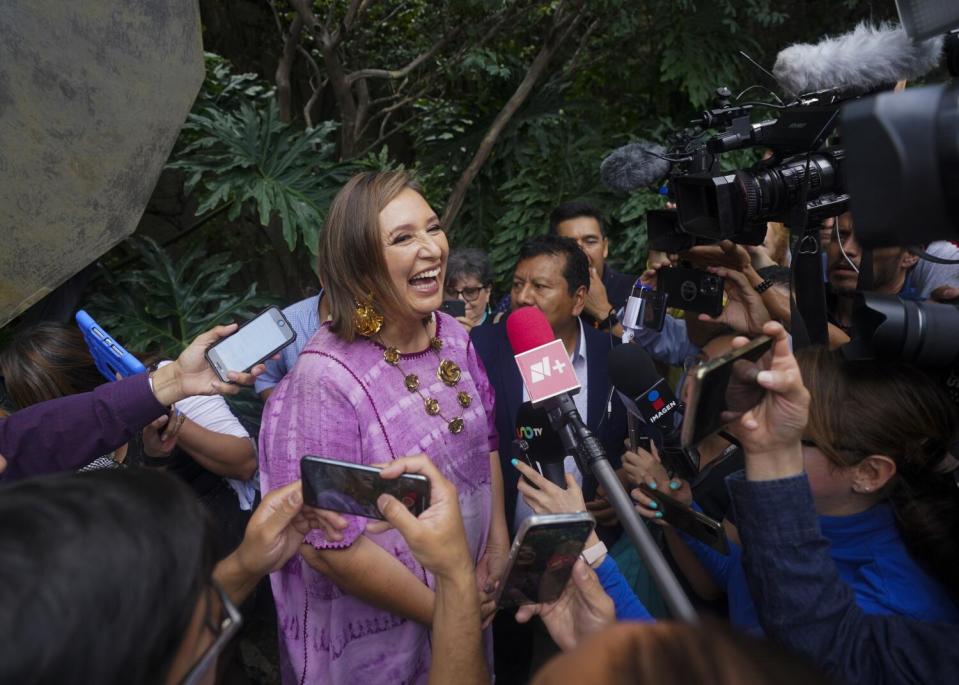 A woman speaks while smiling broadly as microphones are thrust toward her.