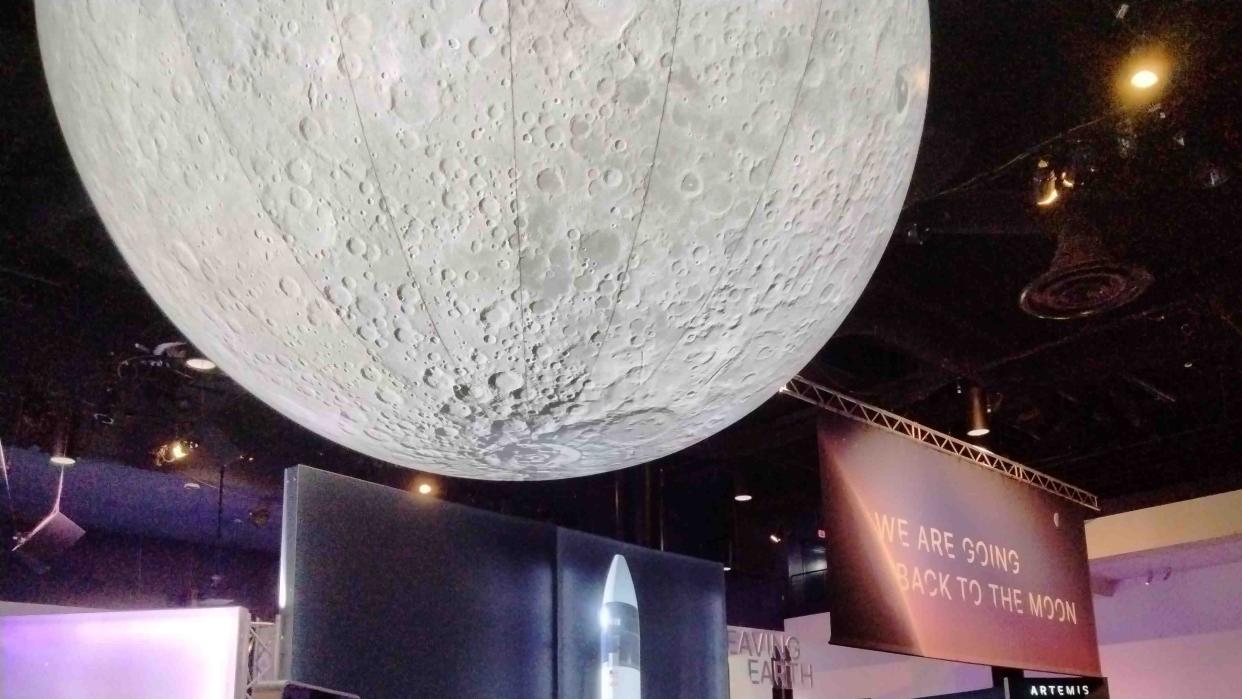  huge moon is attached to the ceiling of a museum. to the right is a big display saying "We are going back to the moon" 