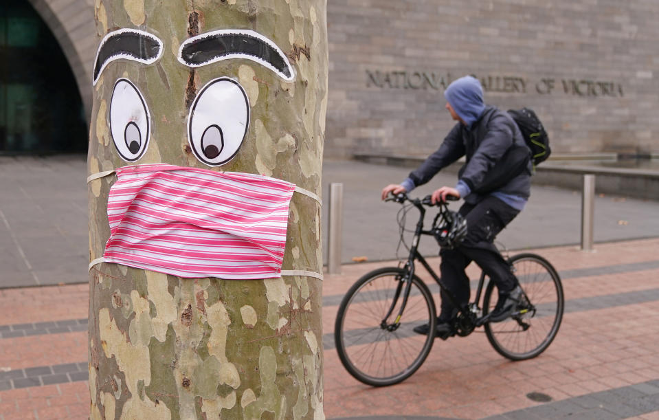 A man rides past a tree with a face mask and eyes stapled to it in Melbourne, Sunday, June 21, 2020. 