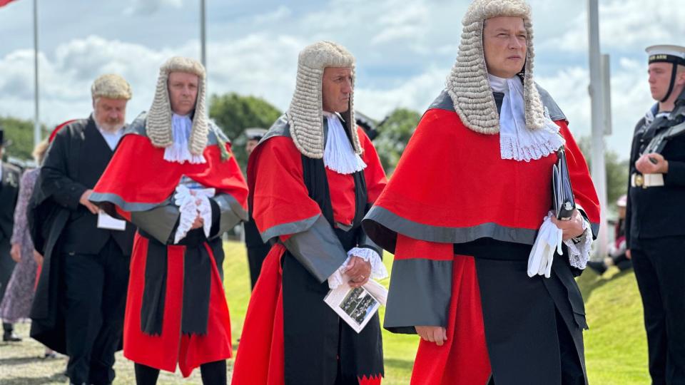 Three men in red robes and judicial wigs and the Attorney General wearing black and a wig behind