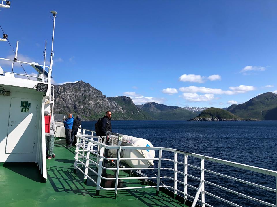 Men in jackets on a ferry deck, with mountains in the background.