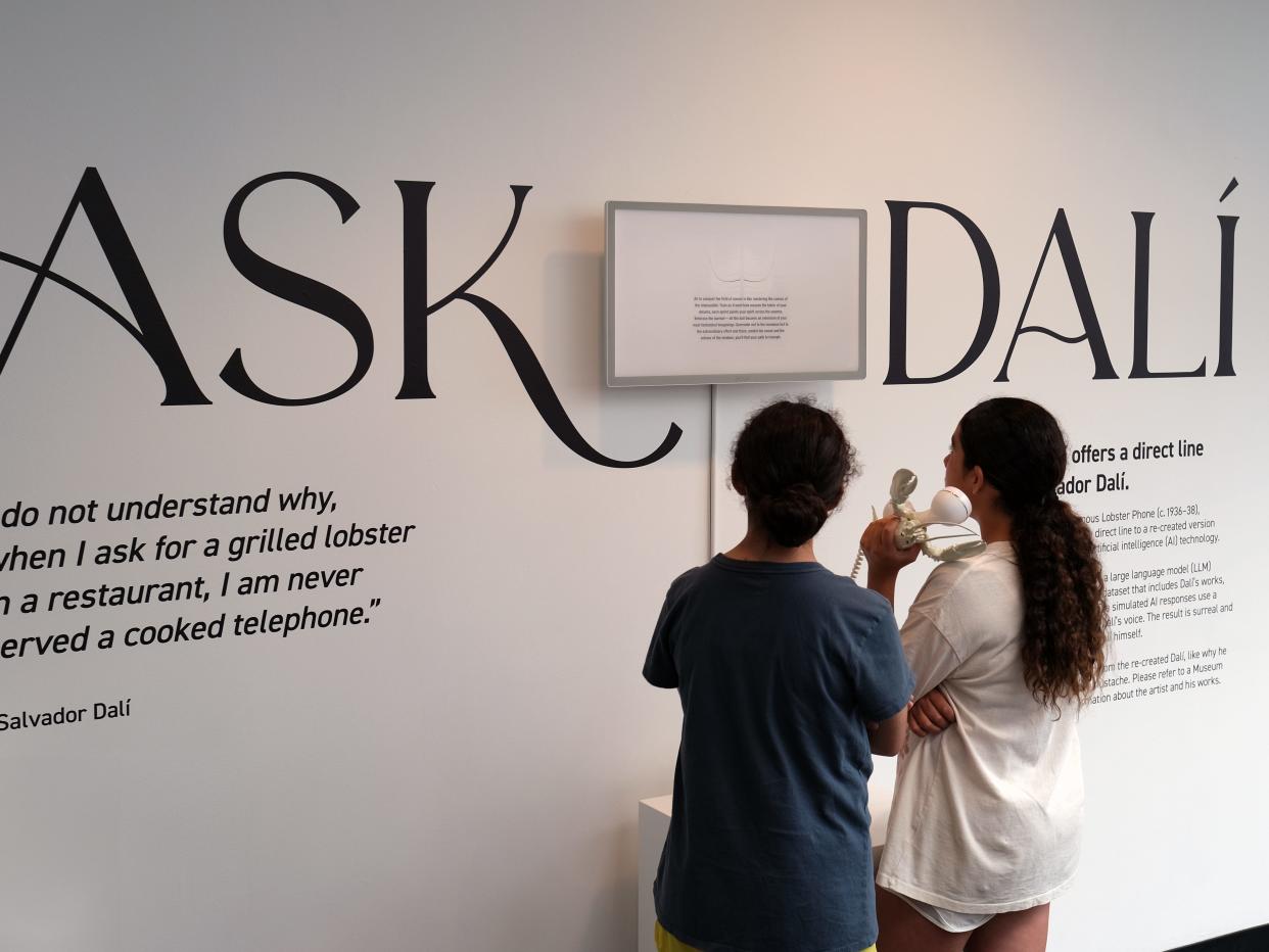 Two girls using a white phone with a lobster staring at a large white wall with the words "Ask Dali"