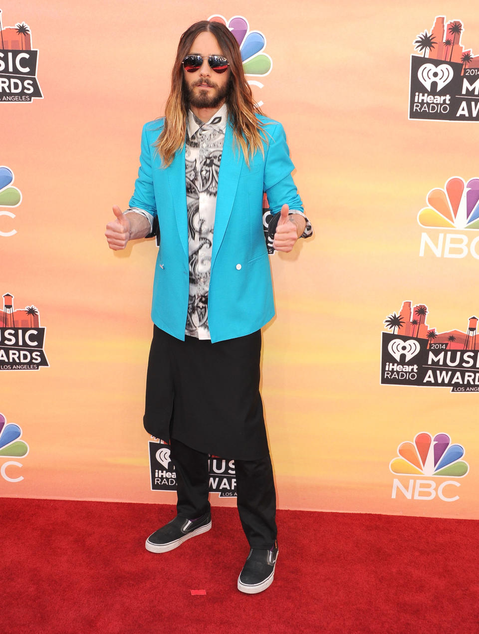 Jared Leto giving thumbs up on red carpet while wearing a skirt over black pants and sand shoes with blue overcoat over white shirt with black patterns, sunset pink/orange promotional wall in background featuring NBC logo