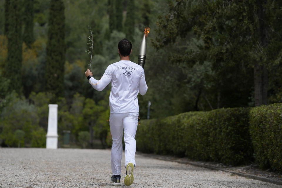 The first torch bearer, Greek olympic gold medalist Stefanos Douskos, runs towards the monument to Pierre de Coubertin, in the background, after the official ceremony of the flame lighting for the Paris Olympics, at the Ancient Olympia site, Greece, Tuesday, April 16, 2024. The flame will be carried through Greece for 11 days before being handed over to Paris organizers on April 26. (AP Photo/Thanassis Stavrakis)