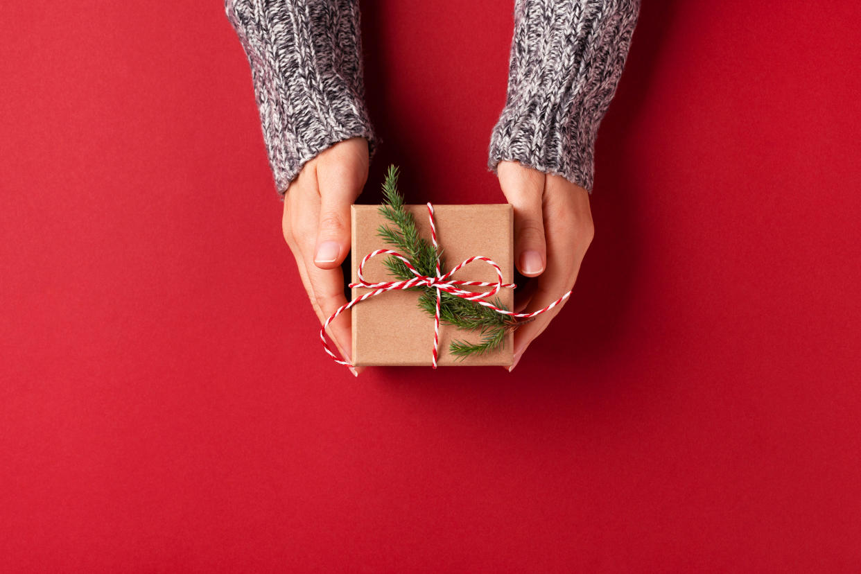 Female's hands in pullover holding Christmas gift box decorated with evergreen branch on red background. Christmas and New Year concept.