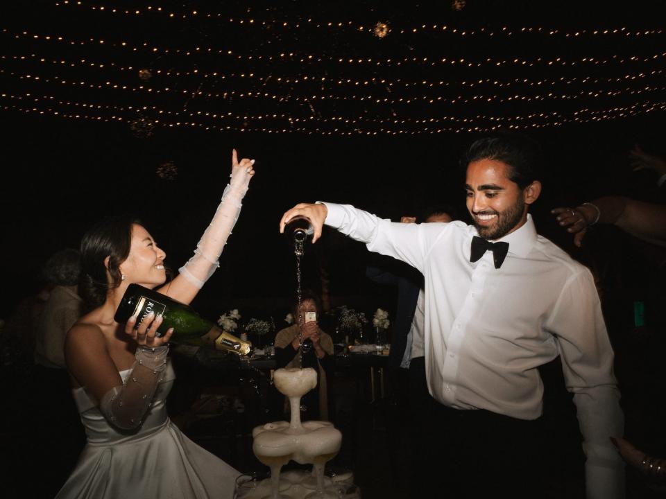 A couple pours champagne into a tower of glasses in front of twinkly lights at their wedding.