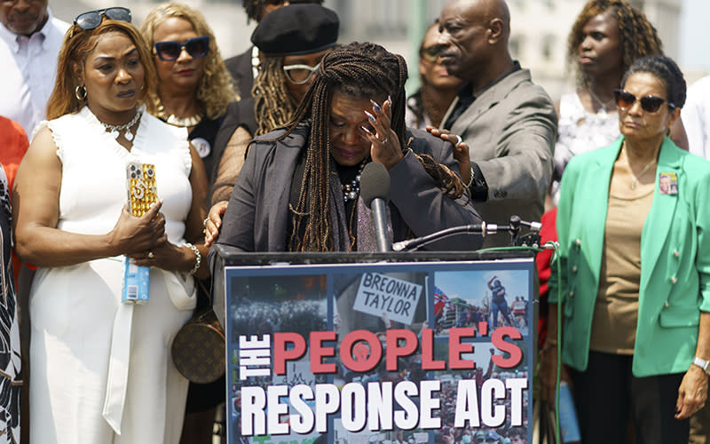 Rep. Cori Bush (D-Mo.) wipes her eyes during a press conference. She is standing at a podium that displays a sign that reads "THE PEOPLE'S RESPONSE ACT" and shows photos from protests. Behind her is a group of people, two of whom are reaching forward to each rest a hand on her shoulder and arm