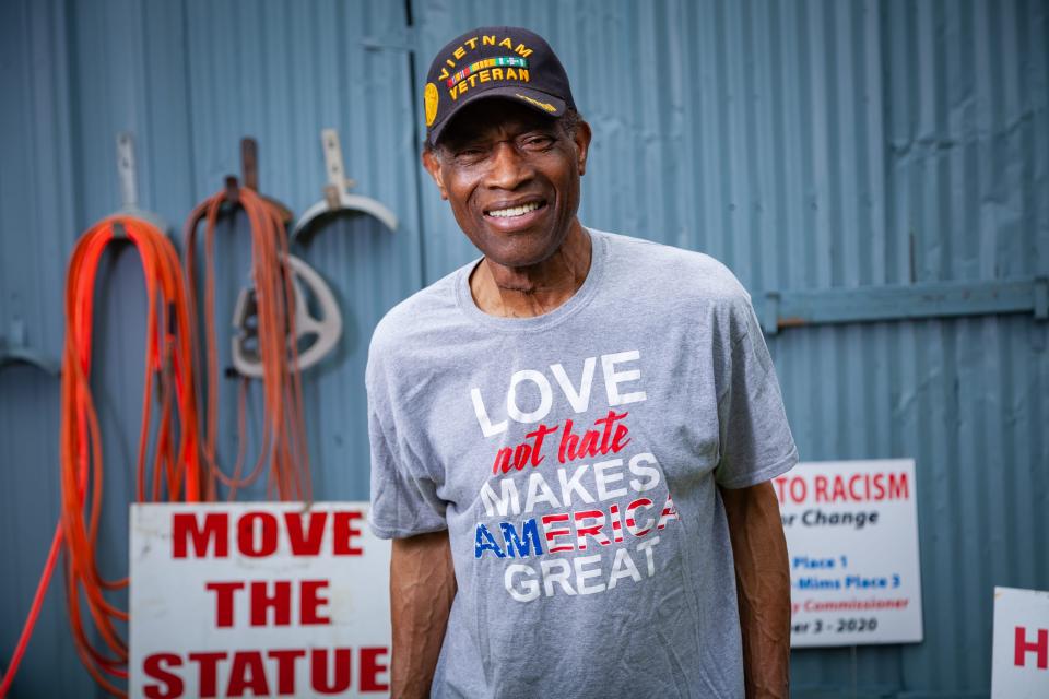 A man stands in front of a shed surrounded by signs like "Move the Statue"