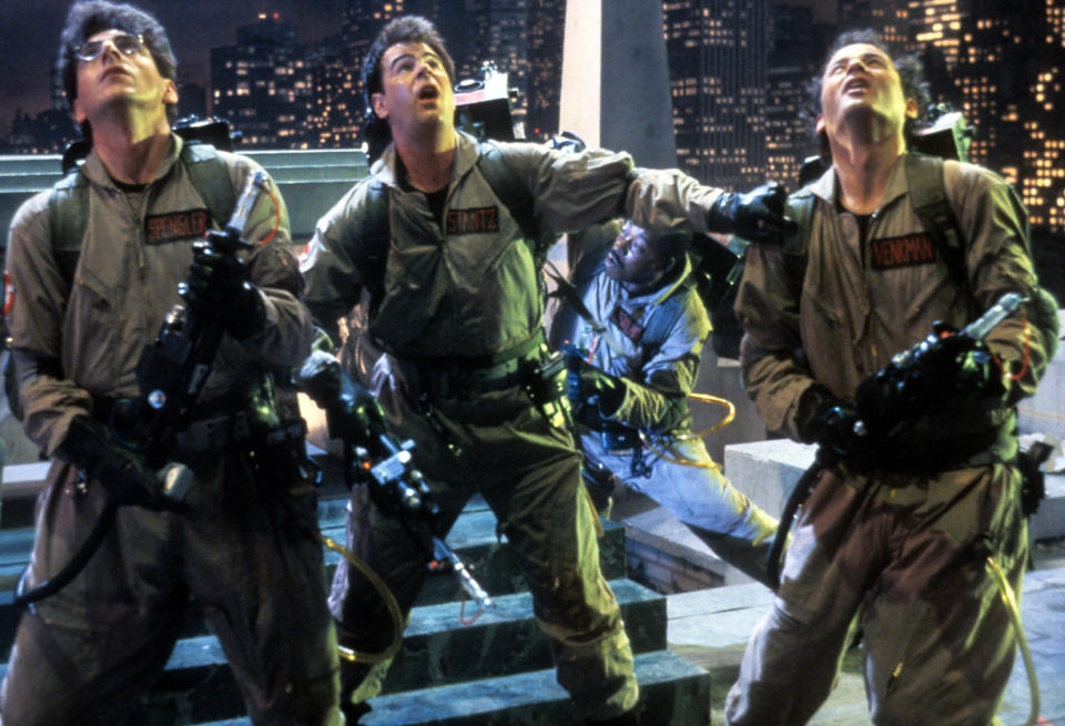 Dan Aykroyd, Bill Murray, Harold Ramis, and Ernie Hudson dressed as Ghostbusters, holding proton packs, looking upwards in a cityscape setting