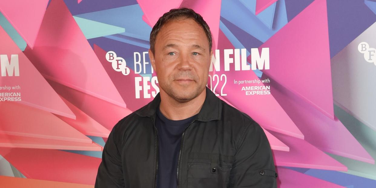 stephen graham stands clasping hands and looking at the camera, he wears black trousers, tshirt, jacket and trainers