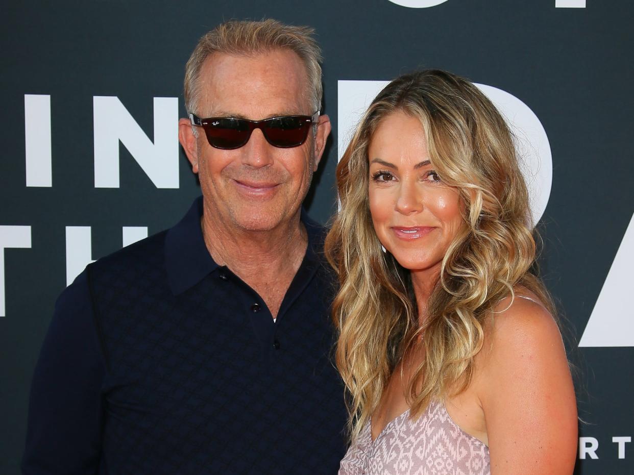 Kevin Costner, who is wearing a black shirt and blacked-out sunglasses, standing with his arm around Christine Baumgartner, who is wearing a sleeveless dress.