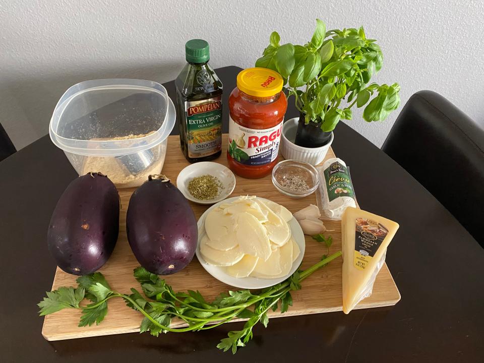 A cutting board filled with ingredients, including eggplant, dried oregano, olive oil, marinara sauce, basil, and cheese.
