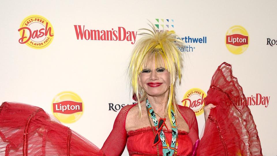 betsey johnson smiles for a photo holding out the ends of her red tulle dress to each side