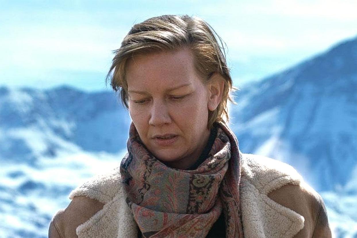 Sandra Hüller wears a heavy coat and scarf and stands in front of mountains covered in snow in a scene from Anatomy of a Fall. 