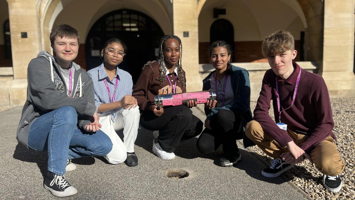 Five students, boys and girls, wear casual clothing and pose for a photo in the school grounds while holding one of the capsules. There is a hole in the pavement in front of them ready for the capsule to be buried in.