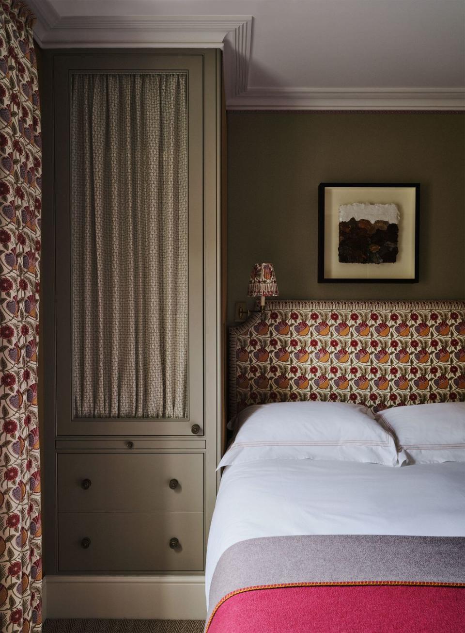 a guest bedroom has matching fabric on the headboard, sconce, and curtains, bed is dressed in white linens and a gray and rose wool blanket, built in closet with drawers, framed art above the headboard