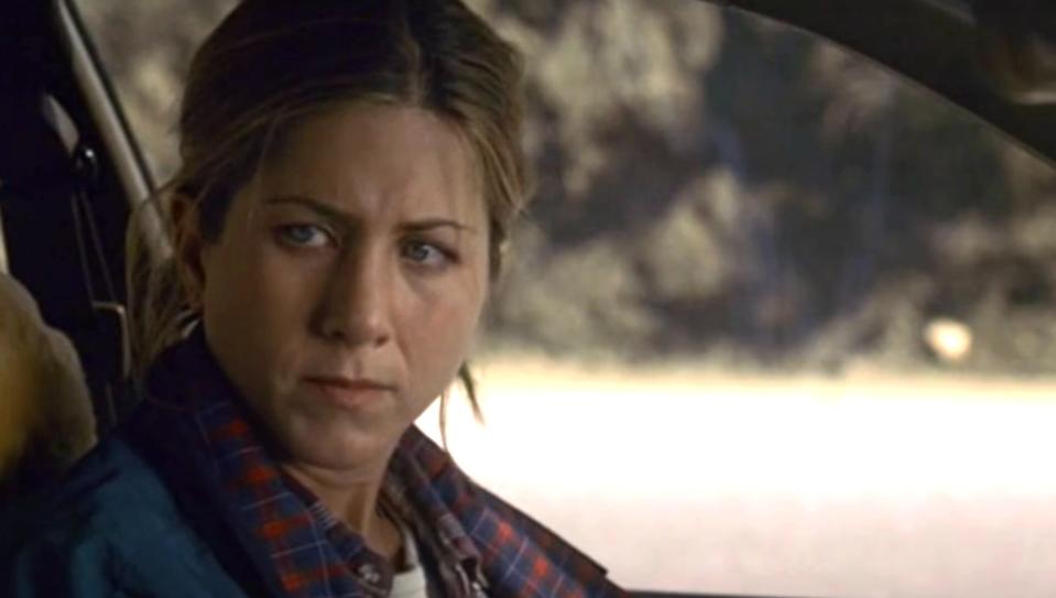 Jennifer Aniston looks concerned while sitting in a car, wearing a plaid shirt with a jacket