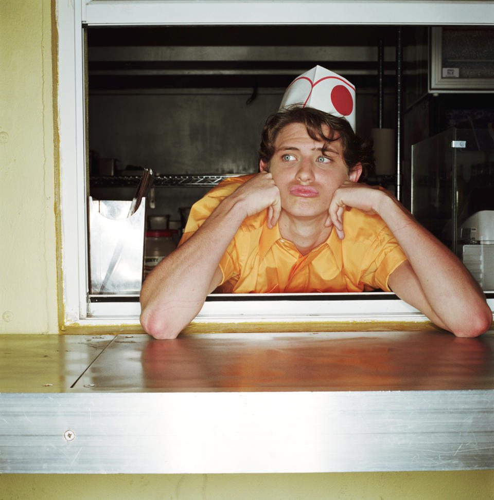 A person in a fast-food worker uniform and hat leans on a counter, looking bored. They are inside a service window, resting their chin on their hands