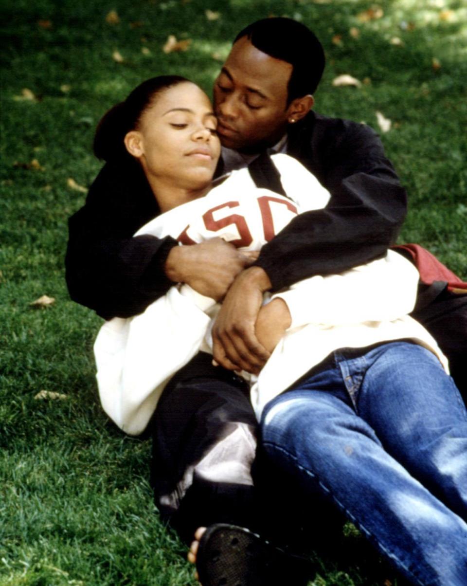 Sanaa Lathan and Omar Epps sit on a lawn, with Omar kissing Sanaa's head while she rests her eyes and leans back into him, both wearing casual clothing