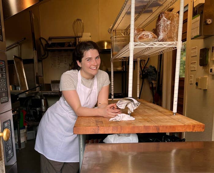 A female chef in an apron and casual pants stands behind a wooden counter in a professional kitchen, smiling