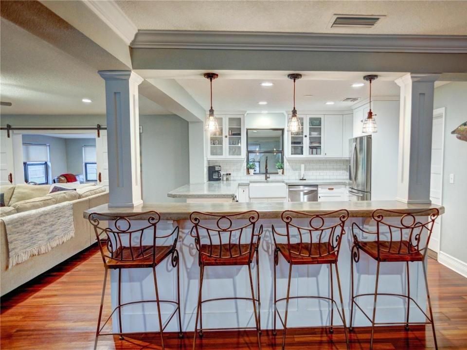 the kitchen with concrete countertops, four stools, white cabinets, and light fixtures next to the living toom