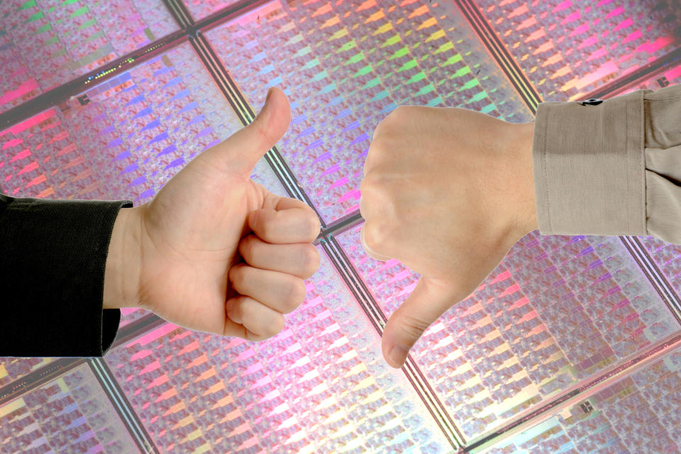 Two hands making thumbs-up and thumbs-down gestures in front of several uncut sheets of semiconductor silicon.