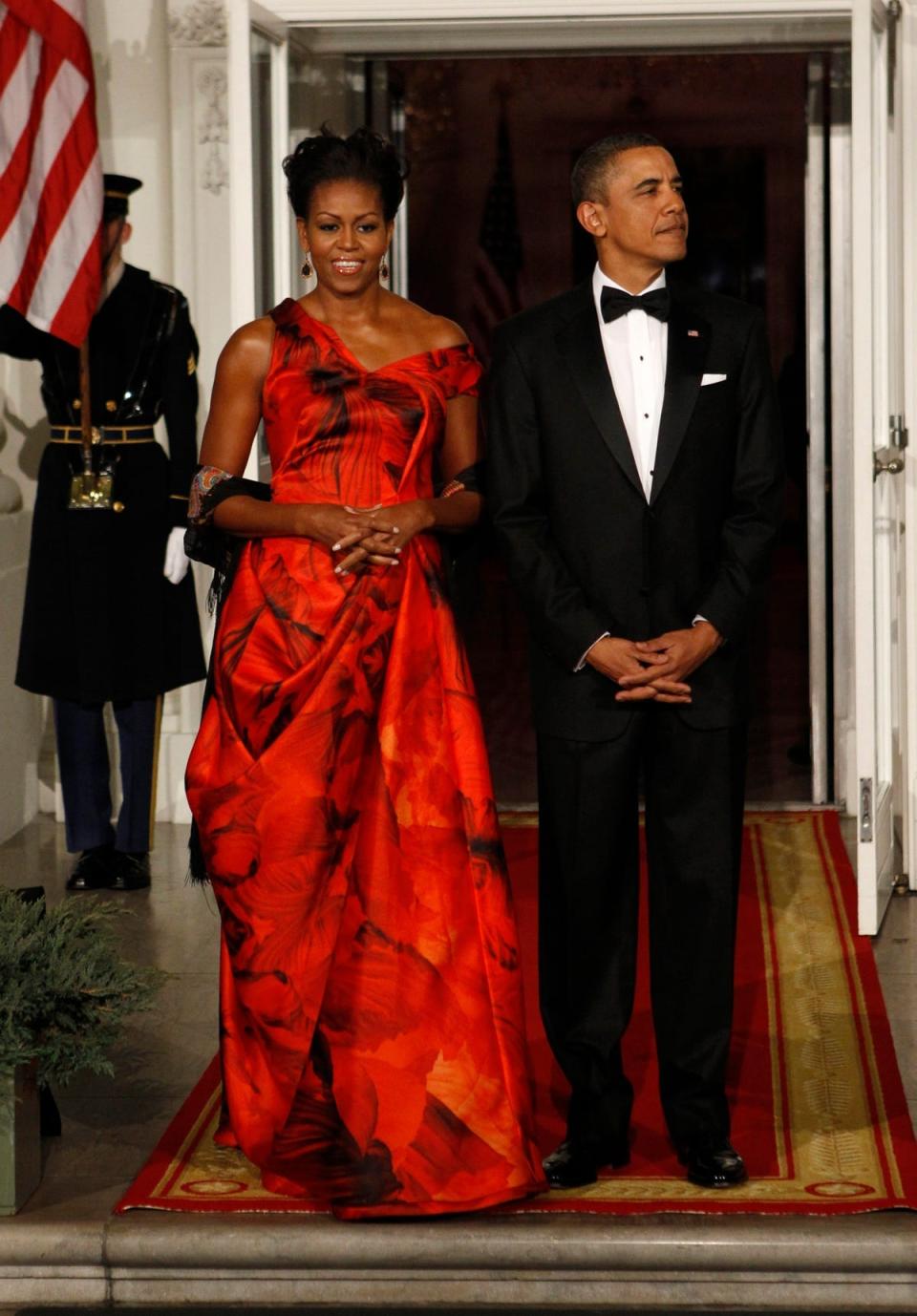 Michelle Obama wears a red Alexander McQueen dress. Barack Obama stands next to her in a tuxedo.