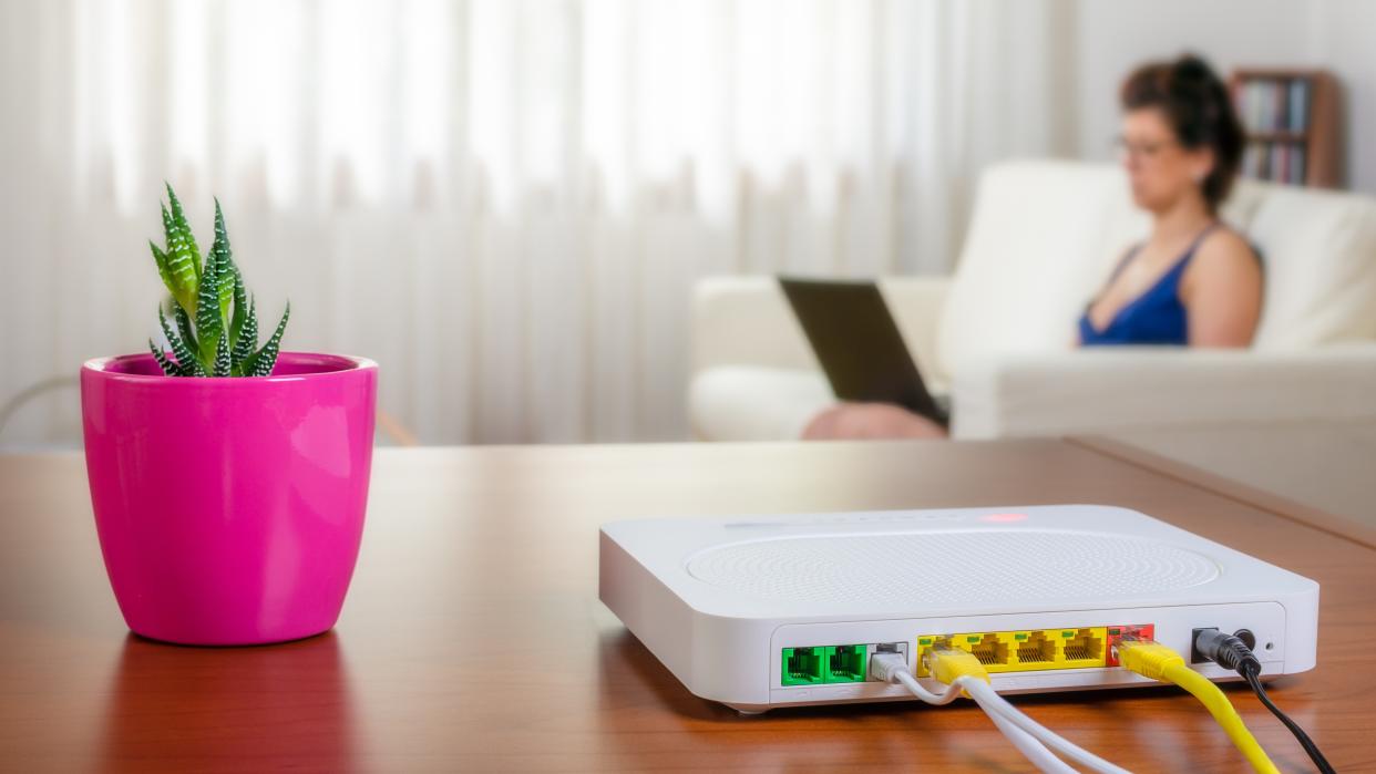  Modem router on a table in a living room. A woman using a laptop while sitting on the sofa is in background. Selective focus. 