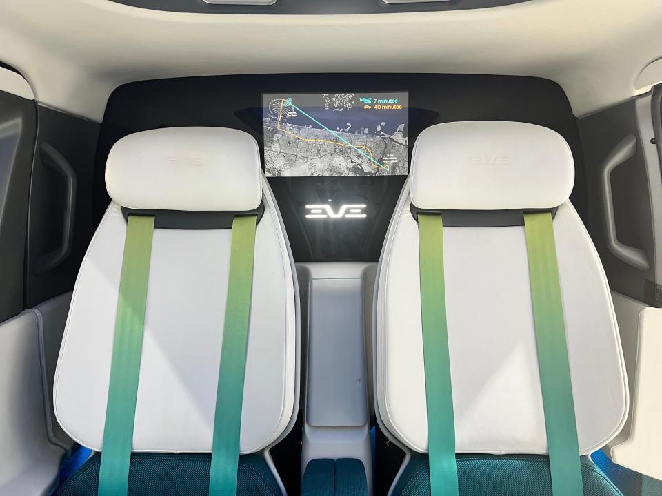The interior of an Eve Air Mobillity eVTOL cabin mockup shows two seats with green-blue seatbelts and a screen on the wall