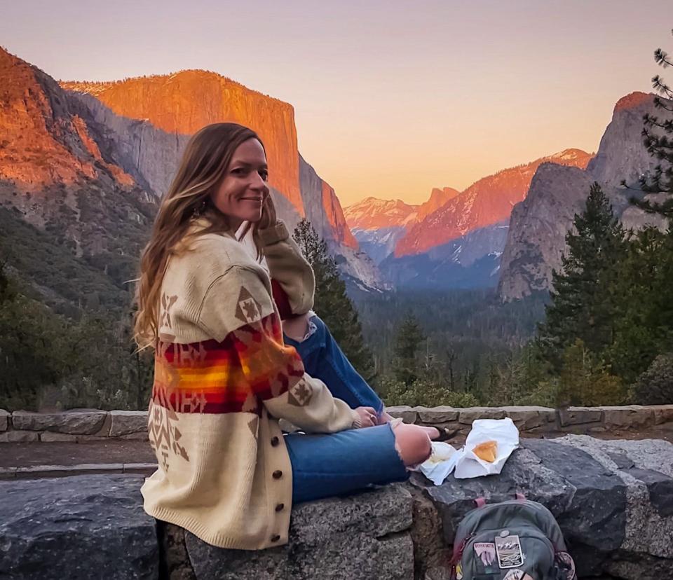 Emily, wearing a sweater and ripped blue jeans, sits on a wall in front of mountains at sunset. Some food and a backpack are placed next to her.