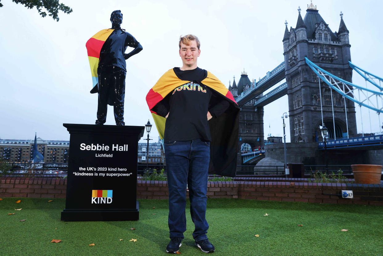 Sebbie Hall stands with his hands on his hips and a colourful cape around his shoulders, in front of his statue in the same pose