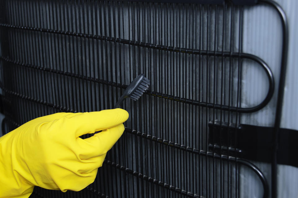 A hand in a yellow glove cleans the fridge radiator grill with a toothbrush close-up.