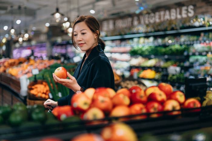 A person, name unknown, stands in a grocery store produce section holding an apple, looking at it thoughtfully. Numerous fruits and vegetables are in the background