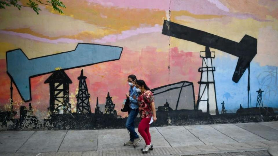 Dos mujeres caminan por la calle en Venezuela junto a un mural en el que hay pintados balancines de petr&#xf3;leo.
