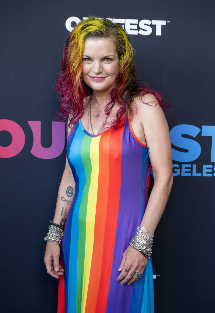 Pauley Perrette stands in front of a backdrop at an event, wearing a rainbow-striped dress with colorful hair and multiple bracelets