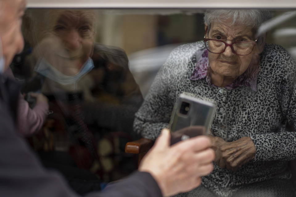 Javier Anto, 90, shows photos of their grandchildren to his wife Carmen Panzano, 92, to combat the ravages of Alzheimer's through the window separating the nursing home from the street in Barcelona, Spain, Wednesday, April 21, 2021. Since the pandemic struck, a glass pane has separated _ and united _ Javier and Carmen for the first prolonged period of their six-decade marriage. Anto has made coming to the street-level window that looks into the nursing home where his wife, since it was closed to visits when COVID-19 struck Spain last spring. (AP Photo/Emilio Morenatti)