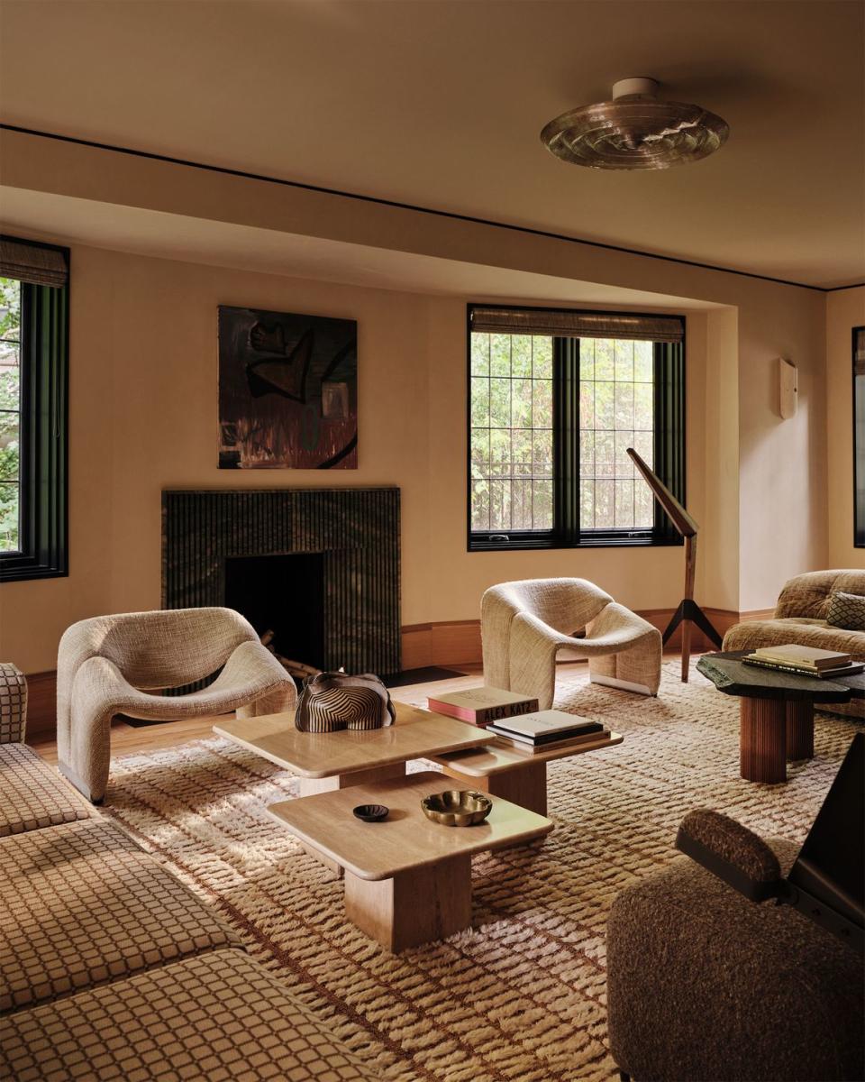 a living room is decorated in tones of brown and beige including two sofas, two modern armchairs, three travertine cocktail tables of different heights, a fireplace, floor lamp, and patterned rug