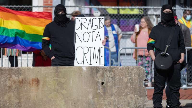 A man at a protest holding a sign that reads drag is not a crime" with a Pride flag in the background.