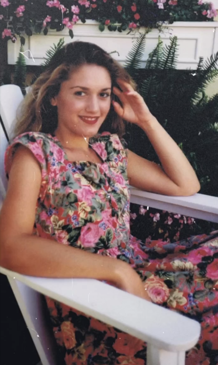 Gwen is sitting in a floral dress, smiling and touching her hair in front of the leaves.