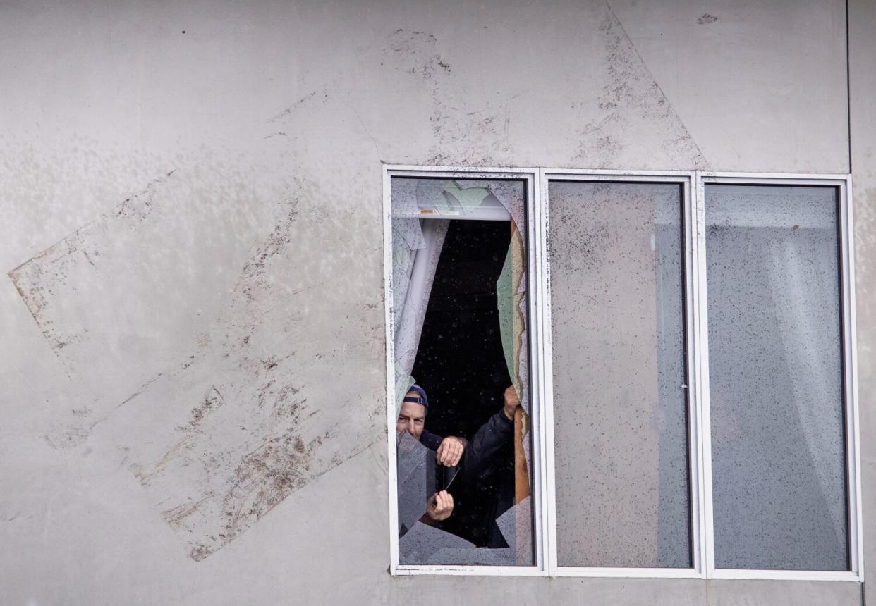 Workers remove broken glass from inside a window, and an imprint made by flying debris is on the wall next to the window.