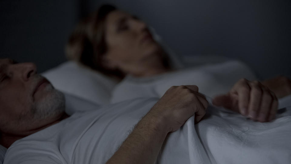 An older couple lies in bed under white sheets, facing away from each other with eyes closed