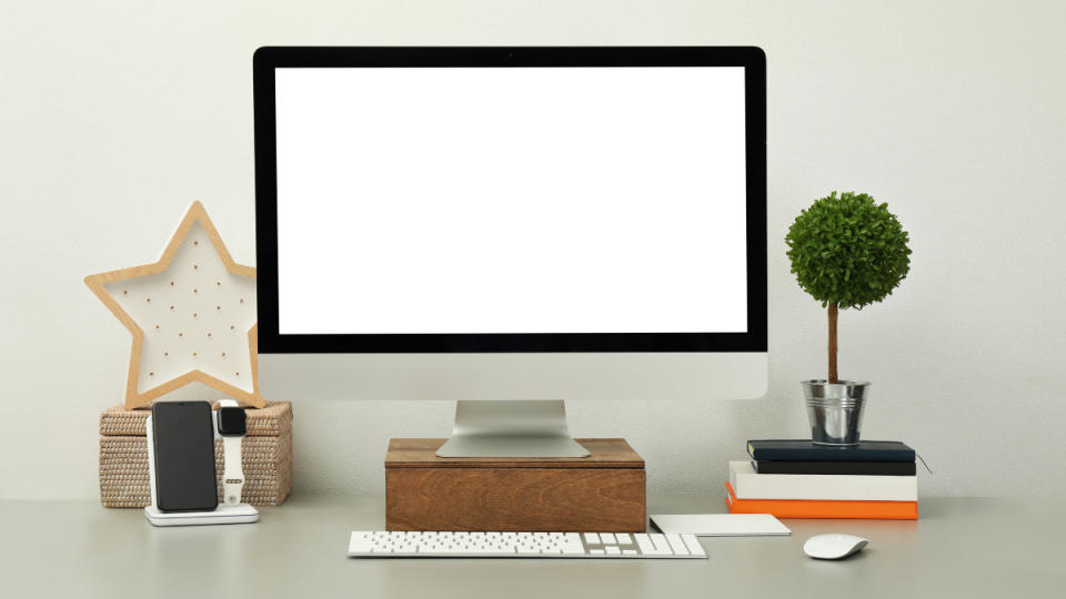 A computer monitor elevated on a desk beside a keyboard, mouse, books and a plant