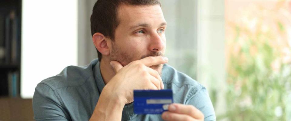 Pensive man holding a credit card thinking looking away through a window sitting in a coffee shop