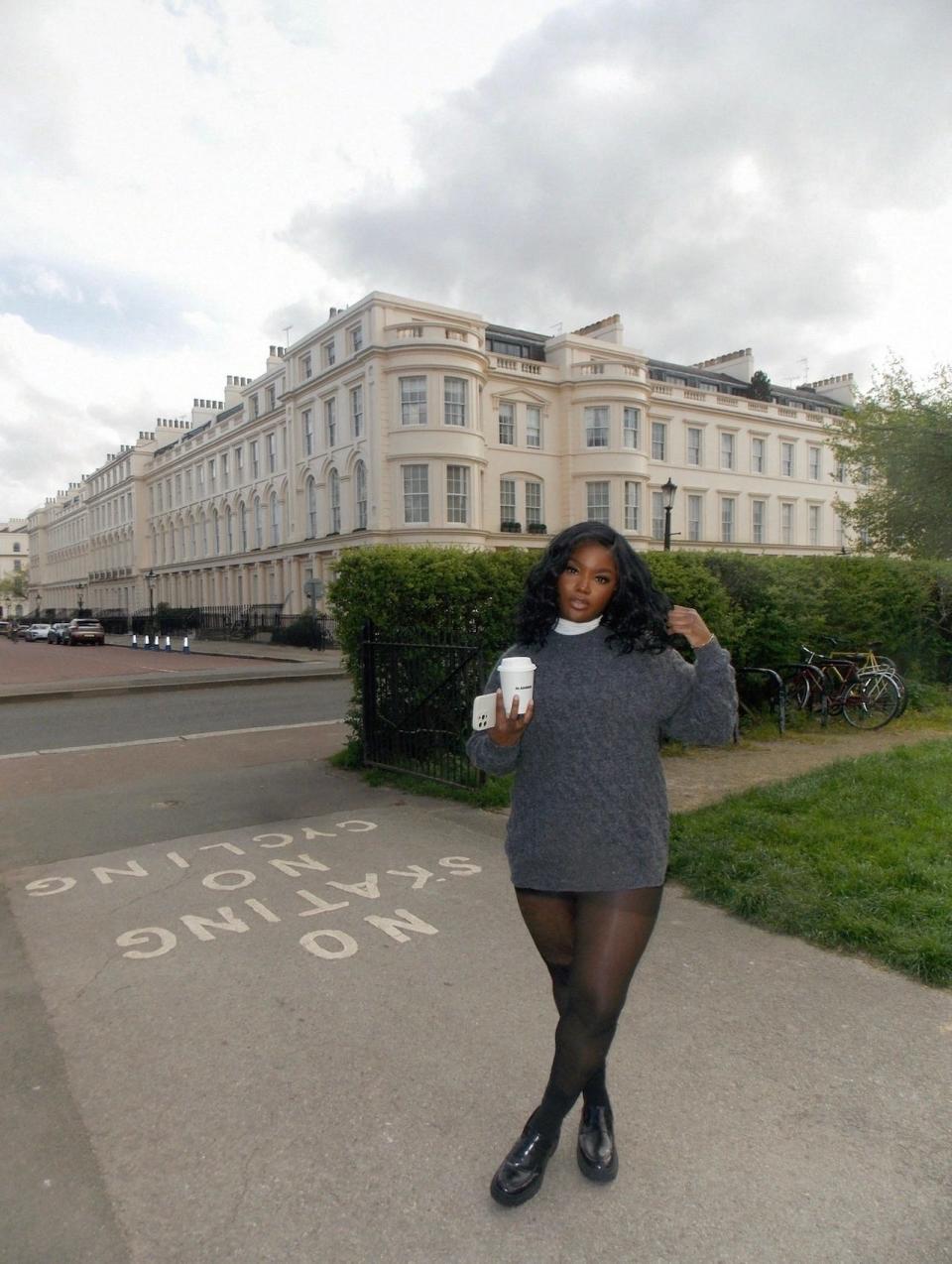 Nkengasong in a blue-gray sweater holding a coffee cup and her cell phone in front of a building and hedges in London.