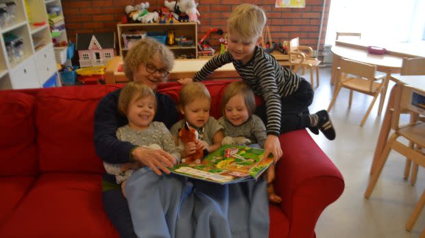Communal grandmother Marjatta Ahonen reads a book to children on a red couch.