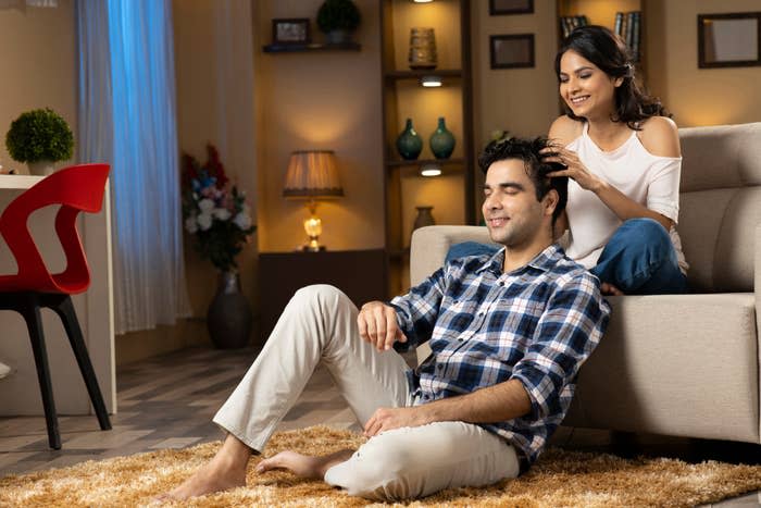 A couple relaxing at home, with the woman affectionately styling the man's hair as they smile