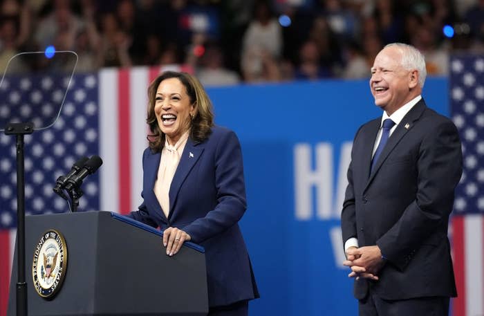 Kamala Harris speaks at a podium with a Vice Presidential seal, smiling, while standing next to Minnesota Governor Tim Walz, who is also smiling